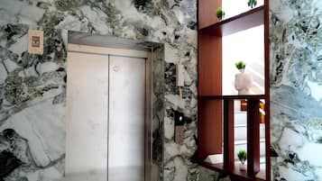 An elevator with a marble wall, a wooden shelf with plants, and a small sign on the wall.