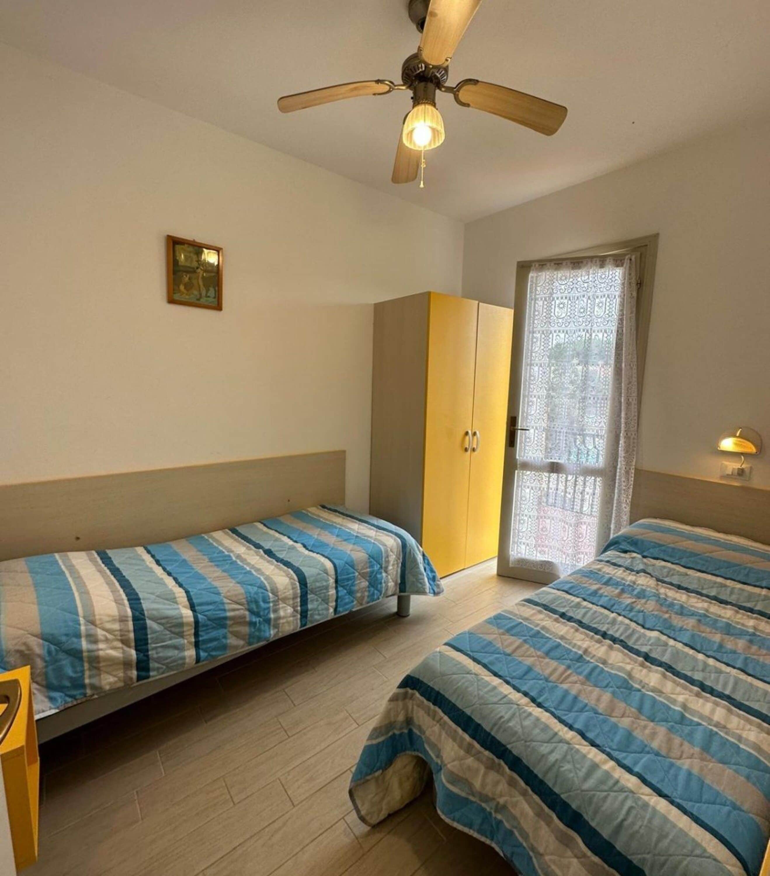 Camera da letto con due letti, una ventola a soffitto, un armadio e una finestra con tende in pizzo.