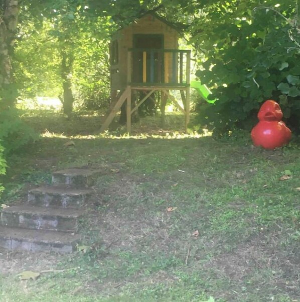 Une maison de jeu en bois entourée de verdure, une sculpture rouge et un escalier en pierre.