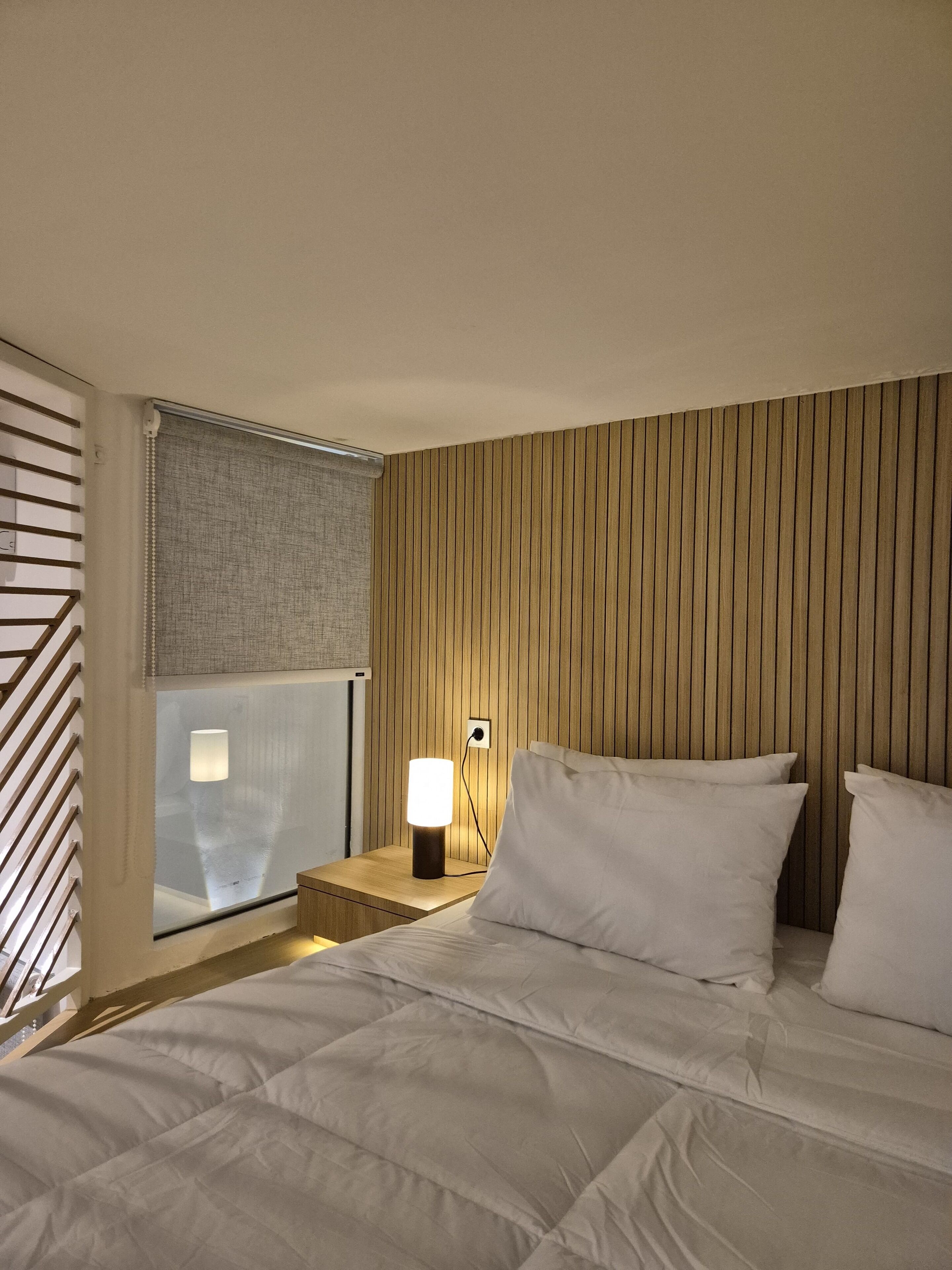 A hotel room with a bed, a nightstand with a lamp, a window with blinds, and a wooden panel wall.