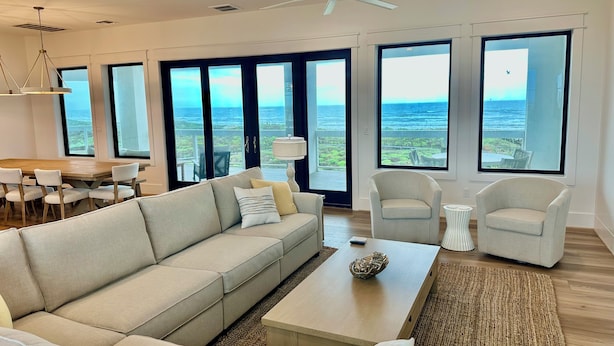 A modern living room with a large sofa, a coffee table, and two armchairs. There are large windows offering a view of the ocean. - Luxury Oceanfront Oasis, Private Pool, Steps to Beach (Port Aransas)