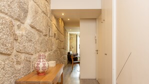 Un mur de pierre, une table en bois avec un vase, et une porte donnant sur une pièce où se trouvent une chaise et une table.