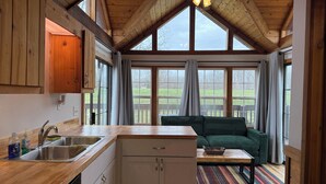 Eine Küche mit Holzarbeitsplatte, Spüle und Schränken. Ein Essbereich mit einem grünen Sofa und einem bunten Teppich. Große Fenster bieten Blick ins Freie.