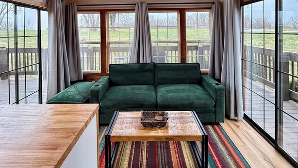 Ein gemütliches Zimmer mit einem grünen Sofa, einem Holztisch und einem Deckenventilator. Große Fenster bieten einen Blick ins Freie.