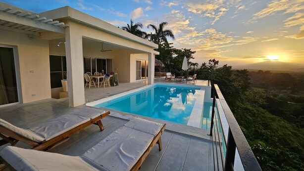 Un área de piscina moderna con sillones de descanso, un porche cubierto y una vista panorámica del atardecer.