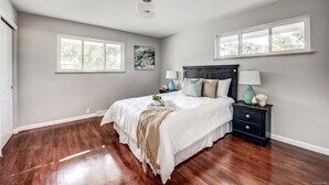 Un dormitorio con una cama grande, dos mesitas de noche, una lámpara de techo y dos ventanas.