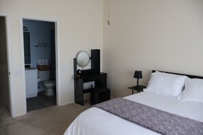 Un dormitorio con cama, mesita de noche, lámpara, cómoda, espejo y baño visible a través de una puerta abierta.