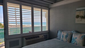 A bedroom with a large window overlooking a beach, a bed with patterned bedding, and a wall-mounted air conditioner. - Bright Beach View Studio • Steps to the Sand & Pool (Panama City Beach)