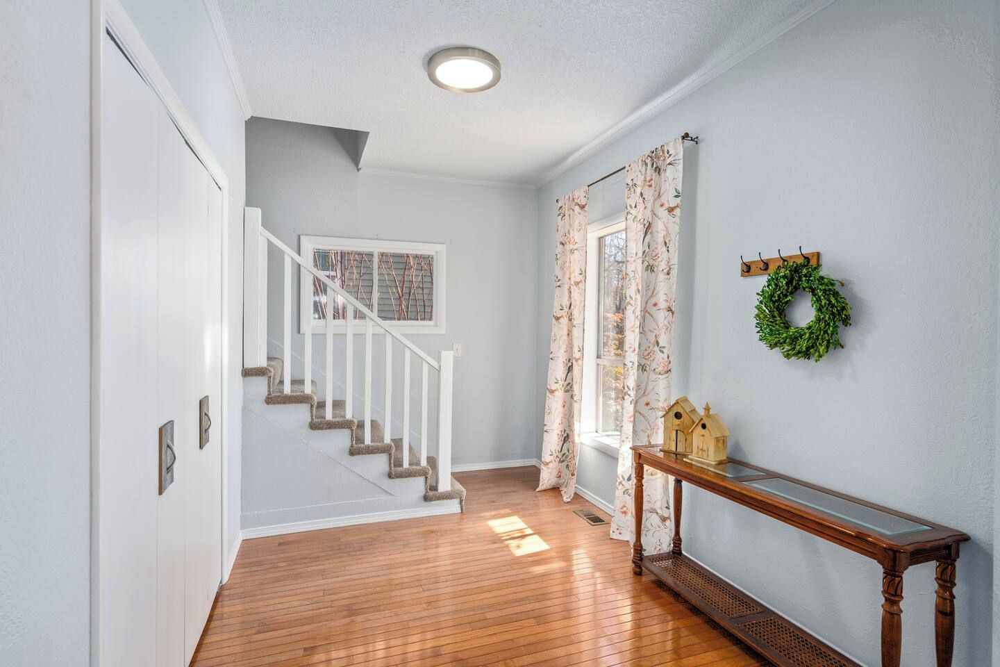 A hallway with a wooden floor, a white staircase, a window with a view, and a small table with a wreath.