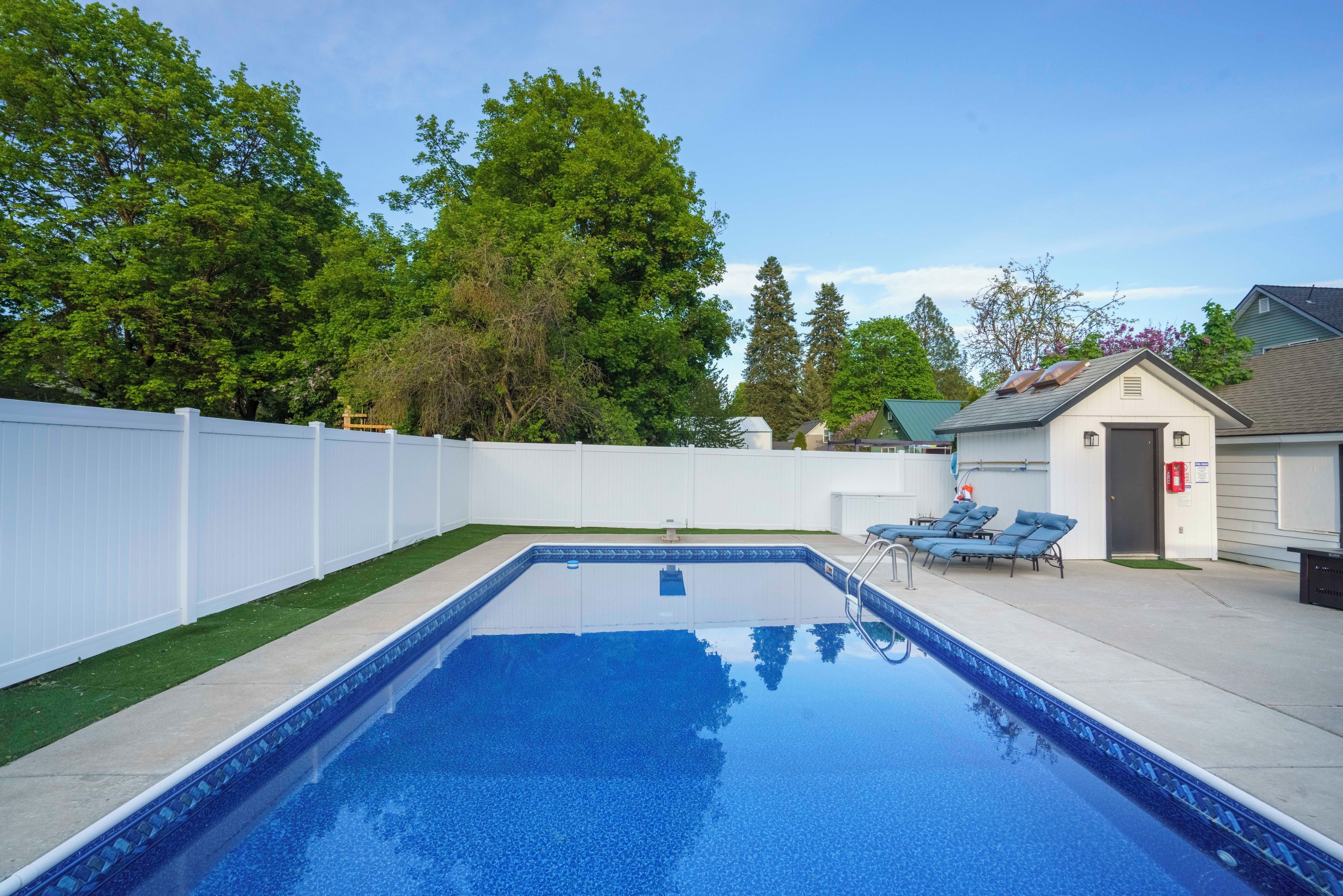 Una piscina rectangular rodeada por una cerca blanca, un pequeño cobertizo y algunas sillas de descanso.