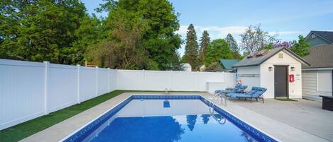 Una piscina rectangular rodeada por una cerca blanca, un pequeño cobertizo y algunas sillas de descanso.