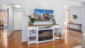 Una sala de estar con chimenea, un televisor montado en un mueble blanco, y vistas de un paisaje urbano a través de la ventana.