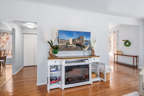 A living room with a fireplace, a television mounted on a white entertainment unit, and a view of a cityscape through the window. - Heated Pool & Hot Tub | Modern Family Retreat (Coeur d'Alene)