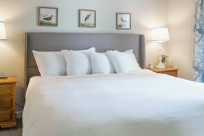 A neatly made bed with white linens, a grey upholstered headboard, and three framed bird paintings above it.