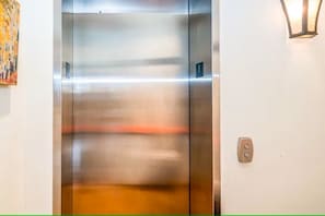 An elevator with a metallic finish, a wall-mounted light fixture, and a door with a number plate.