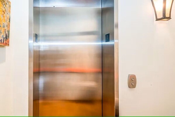 An elevator with a metallic finish, a wall-mounted light fixture, and a door with a number plate.