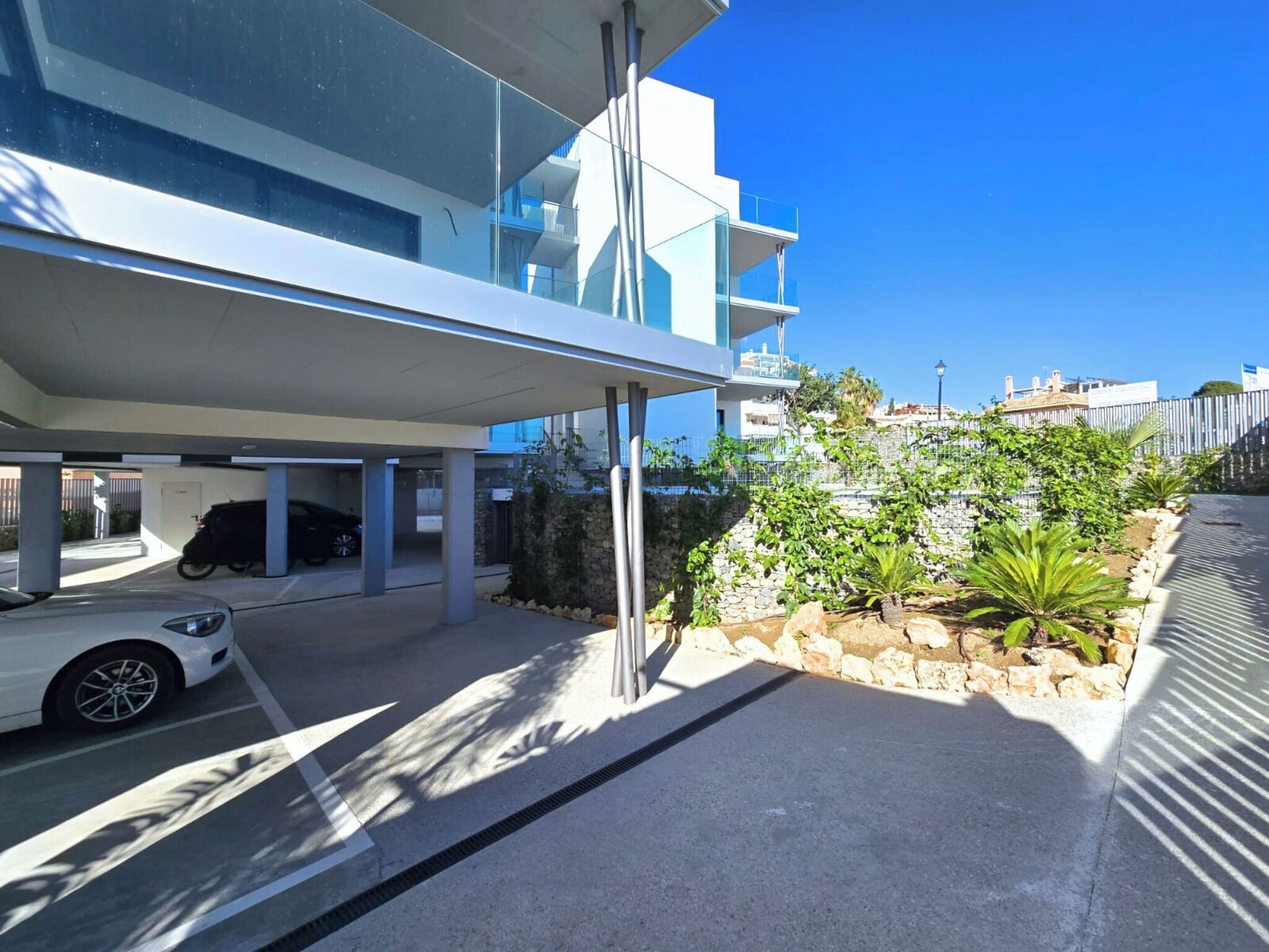 A modern building with a covered parking area, a white car, and a landscaped garden with rocks and plants.