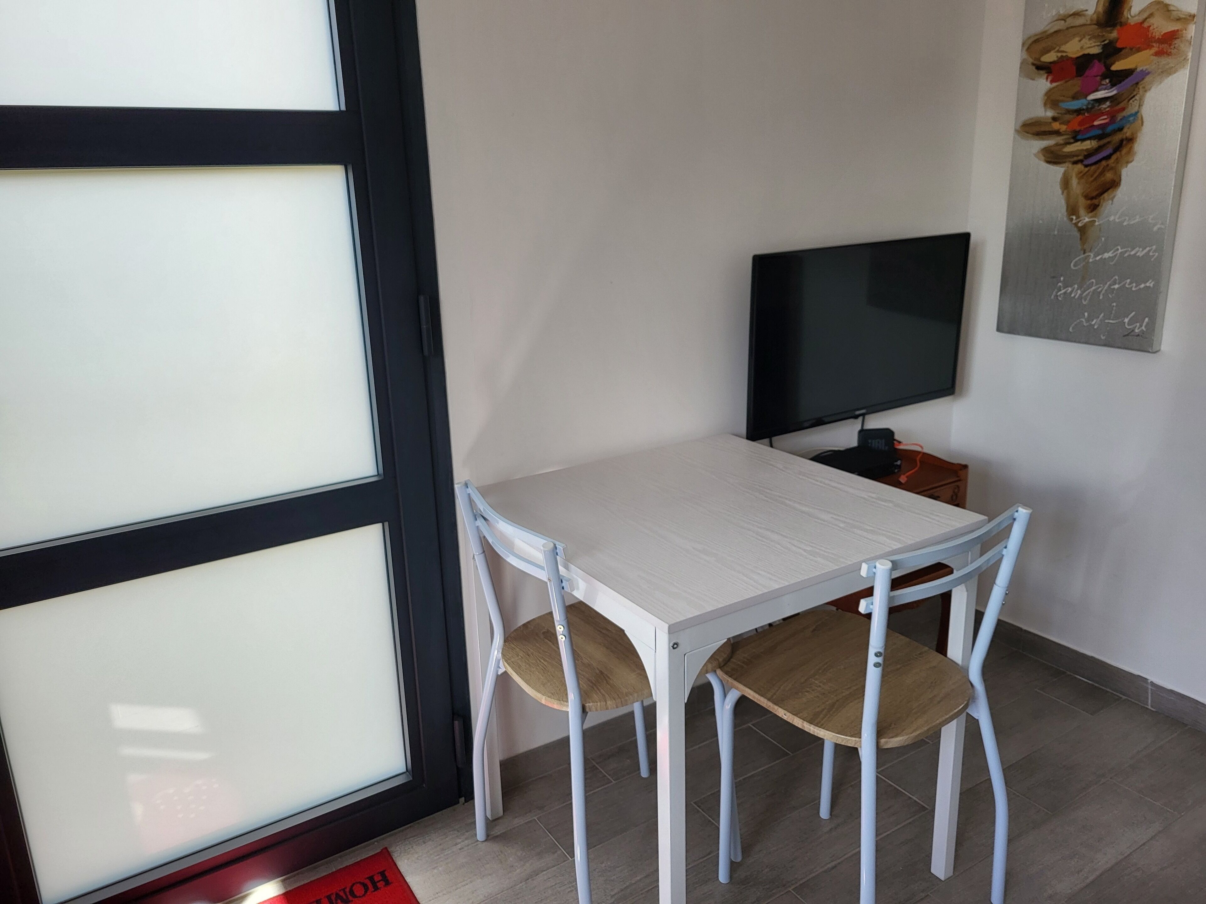 A room with a white table and four chairs, a flat-screen TV on a stand, and a door with frosted glass panels.