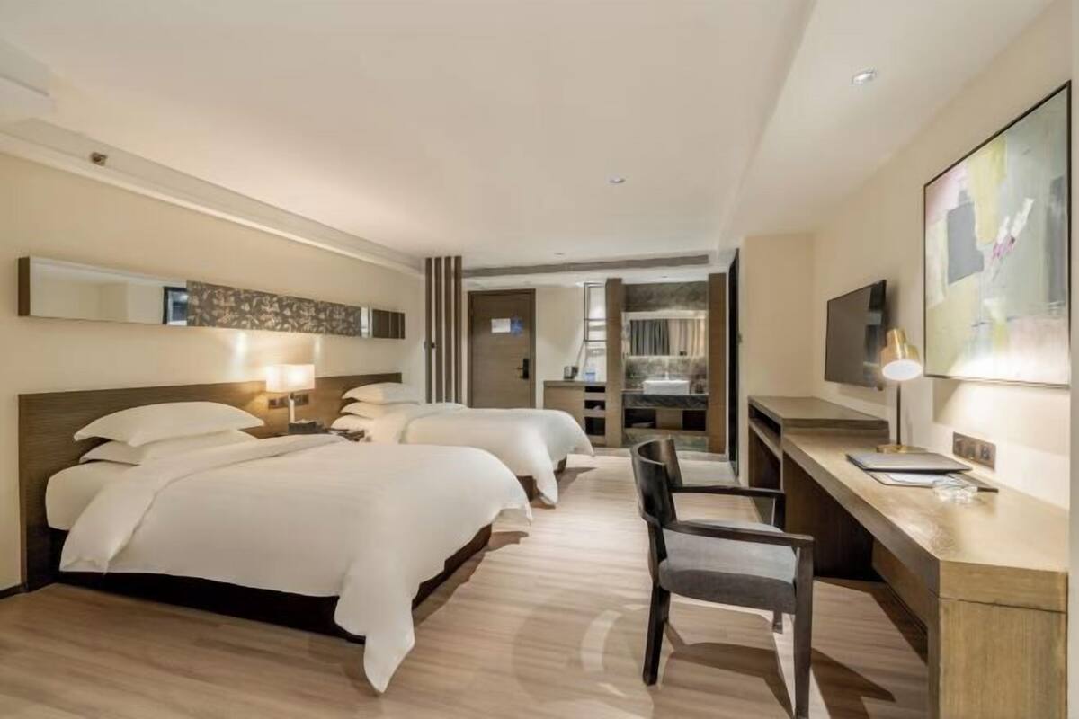 Habitación de hotel con tres camas, un escritorio, una silla, un televisor y un ventanal grande.