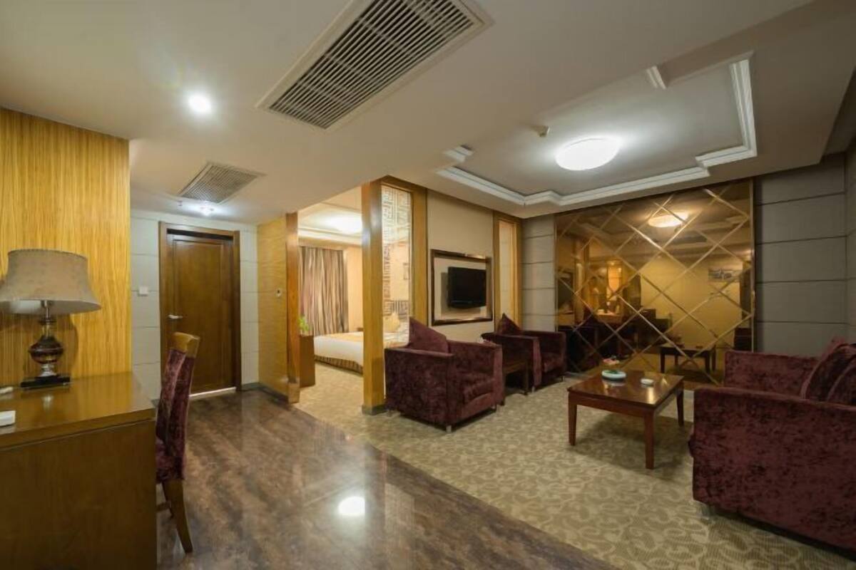 A hotel lobby with a reception desk, seating area with sofas and armchairs, a television mounted on the wall, and a patterned wall design.