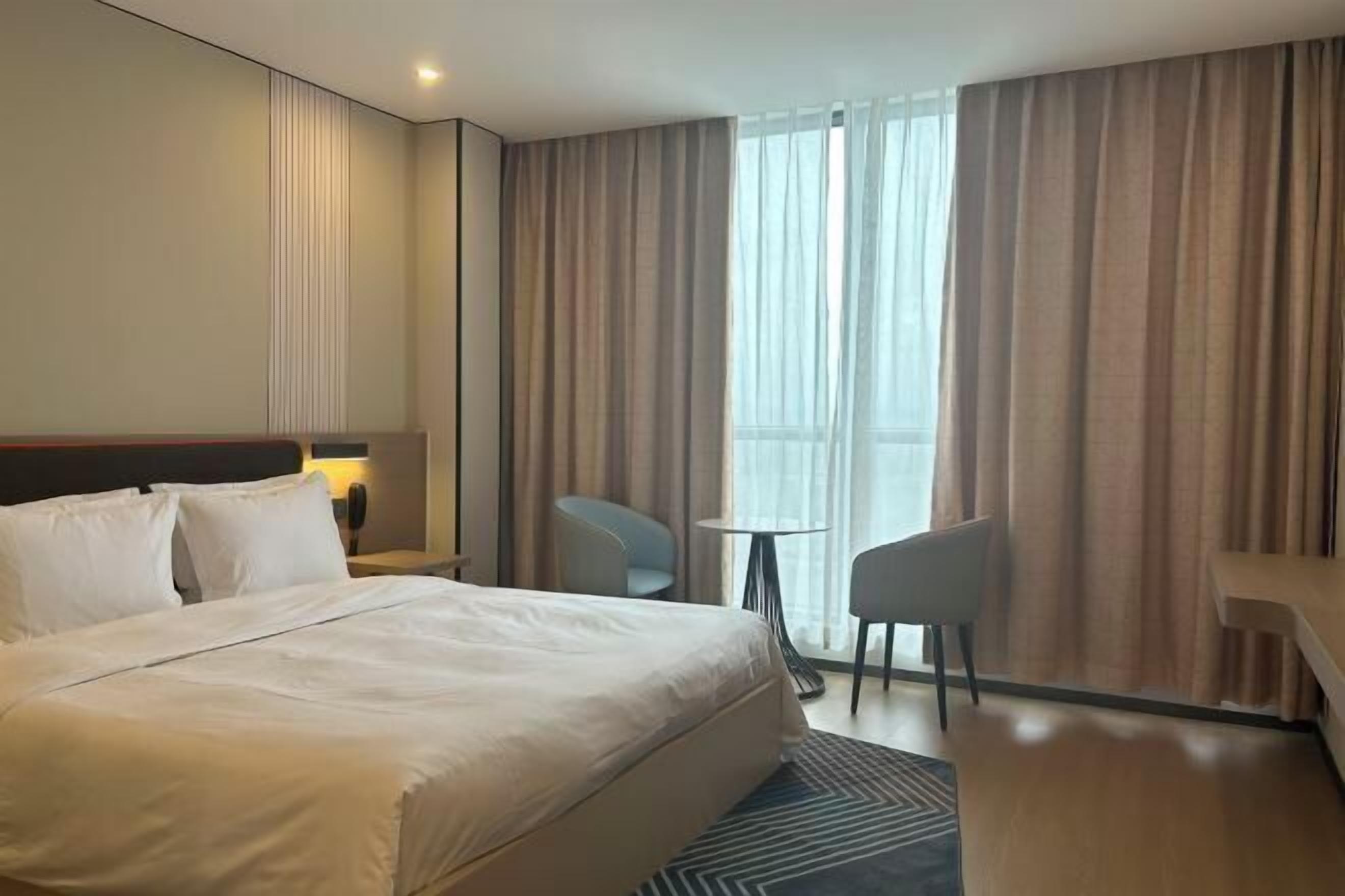 Een hotelkamer met een groot bed, een bureau, twee stoelen, een kleine tafel en een groot raam met gordijnen.