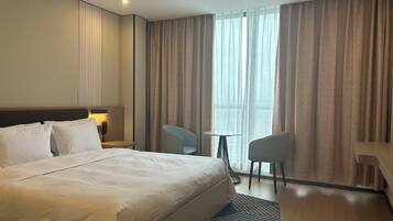 Een hotelkamer met een groot bed, een bureau, twee stoelen, een kleine tafel en een groot raam met gordijnen.