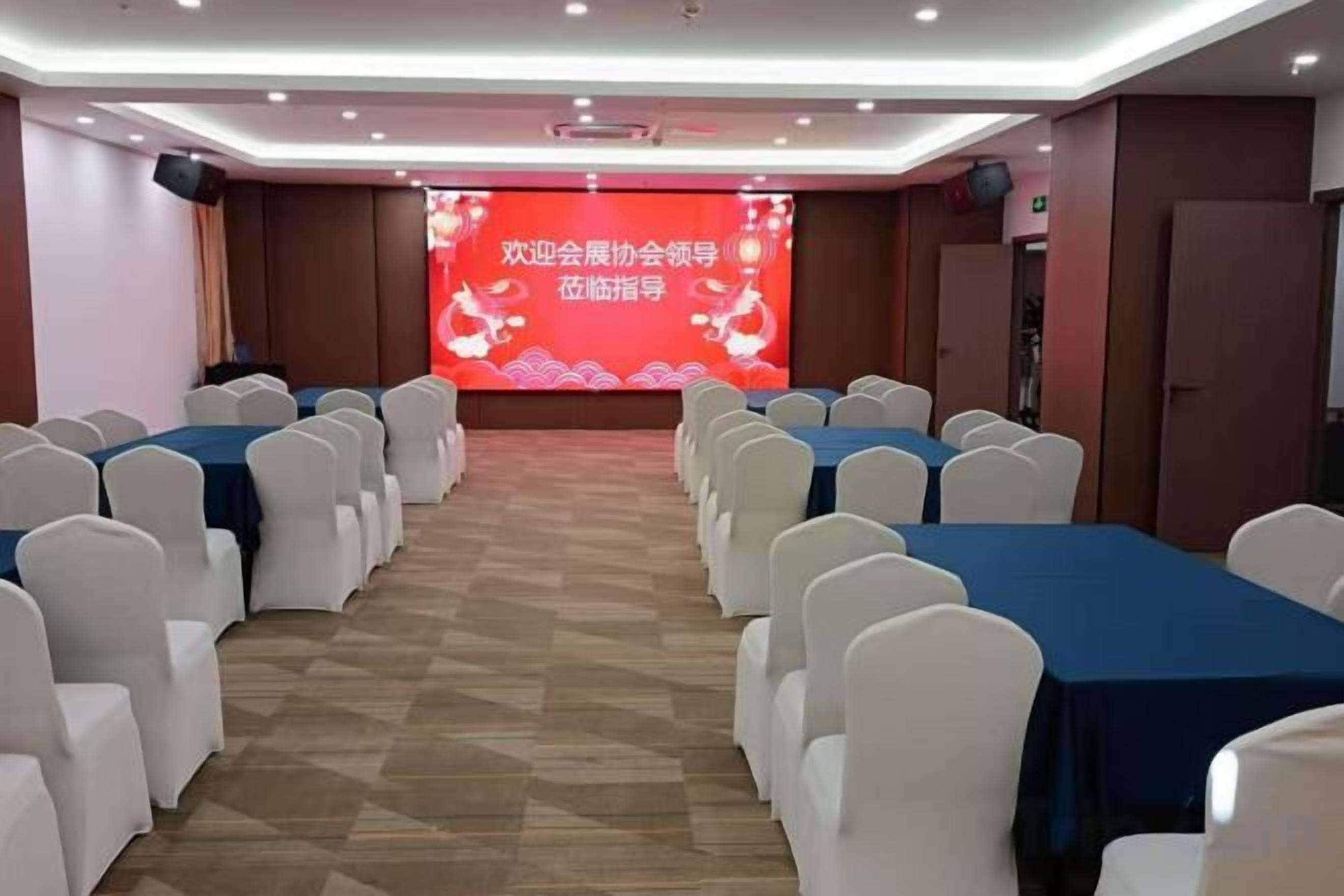 A conference room with rows of white chairs and blue tables, a stage with a red backdrop, and a speaker system.