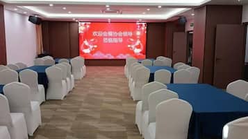 A conference room with rows of white chairs and blue tables, a stage with a red backdrop, and a speaker system.
