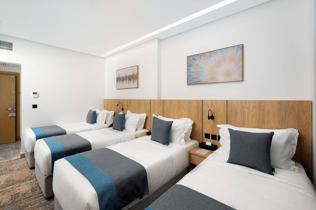 A hotel room with three beds, each with a blue and gray striped bedspread, white pillows, and a wooden headboard. There are two wall-mounted nightstands with lamps and a small table with a tissue box.
