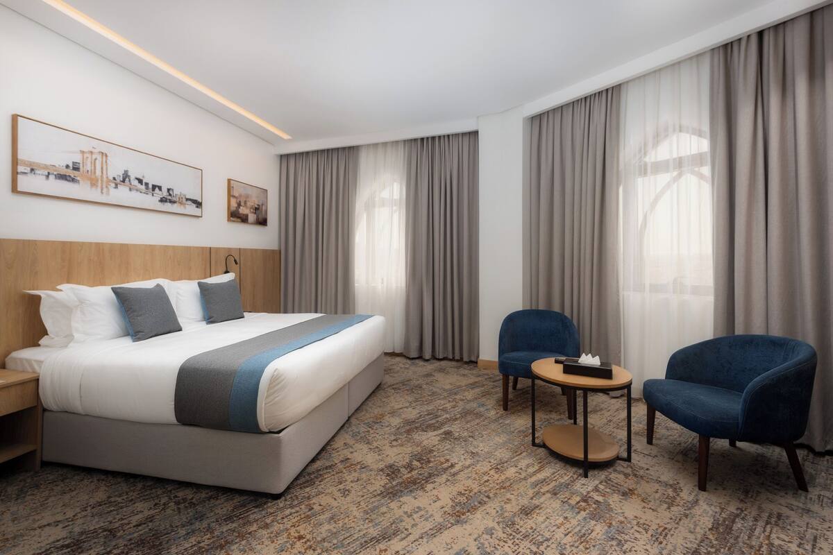 Ett hotellrum med en stor säng, två fåtöljer, ett litet bord och en tavla med en stadsbild på väggen.