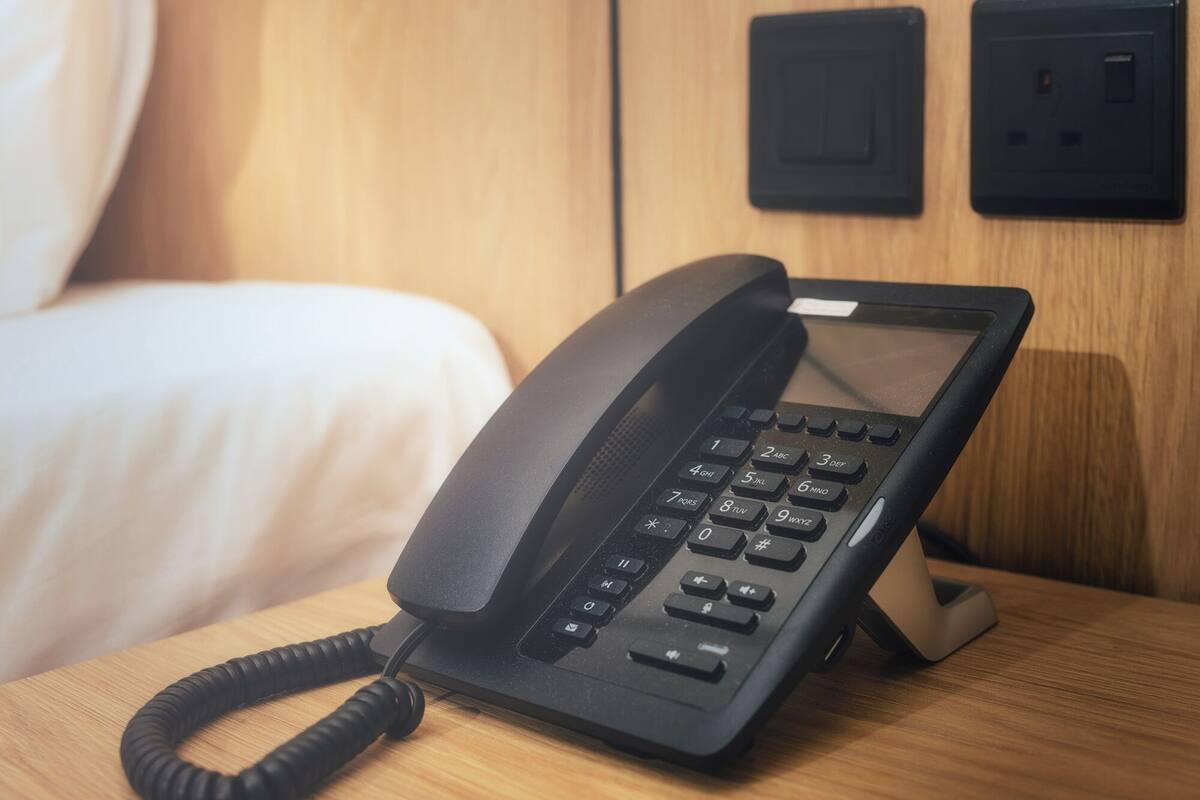 A black telephone on a wooden bedside table in a hotel room.