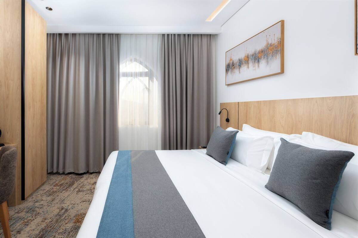 A hotel room with a large bed, a headboard, a bedside lamp, and a window with curtains.