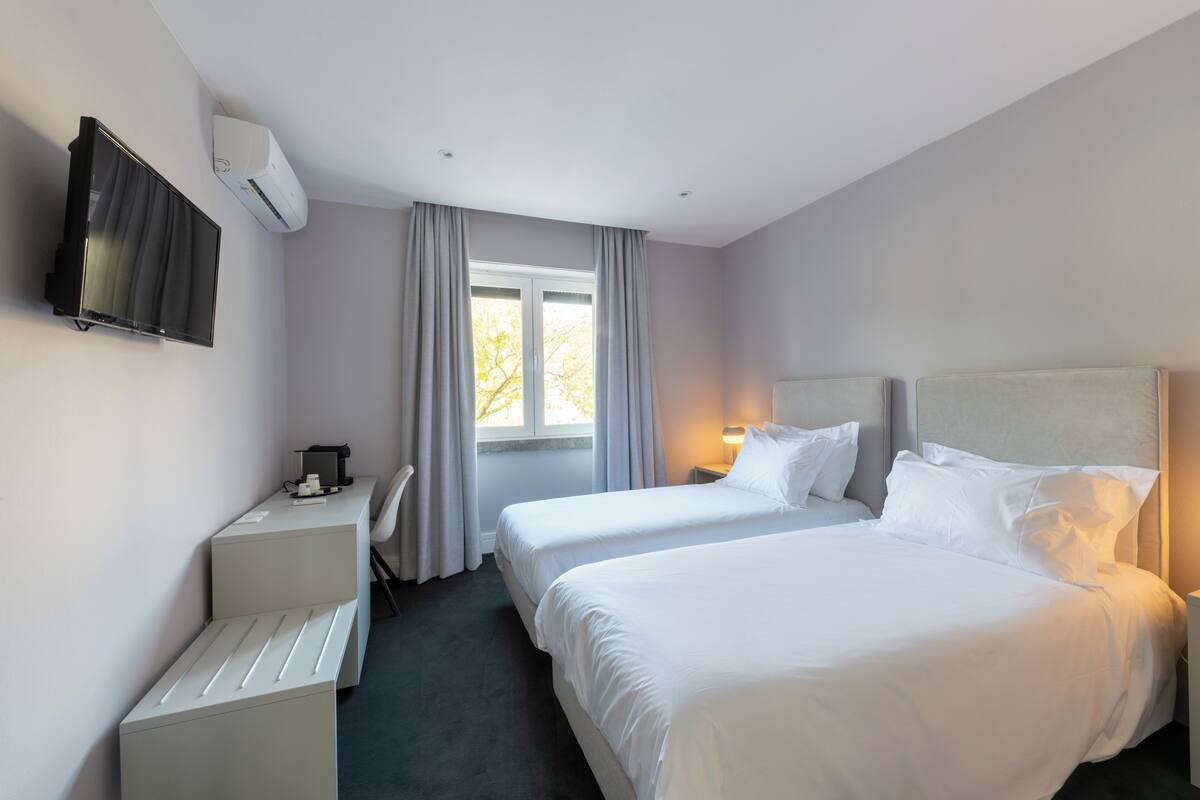 Ein Hotelzimmer mit zwei Betten, einem Schreibtisch, einem Fernseher und einem Fenster mit Vorhängen.