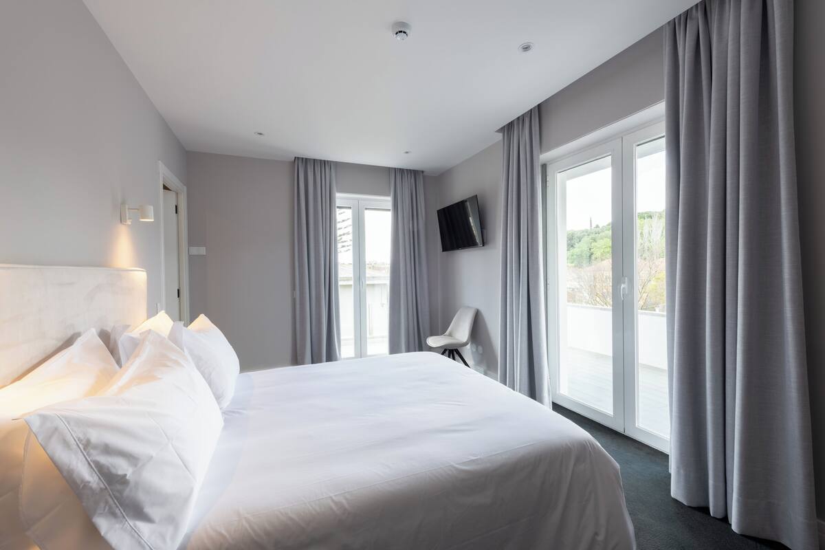 Ein modernes Hotelzimmer mit einem großen Bett, einem Sessel, einem Flachbildfernseher und einem Balkon mit Vorhängen.
