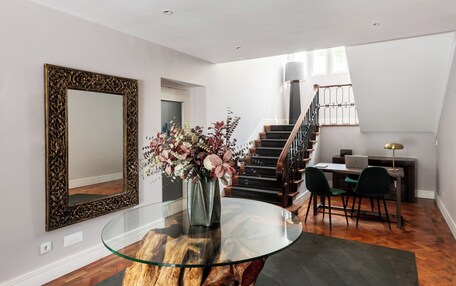 A modern interior with a glass coffee table, a large ornate mirror, a staircase, and a desk with chairs.