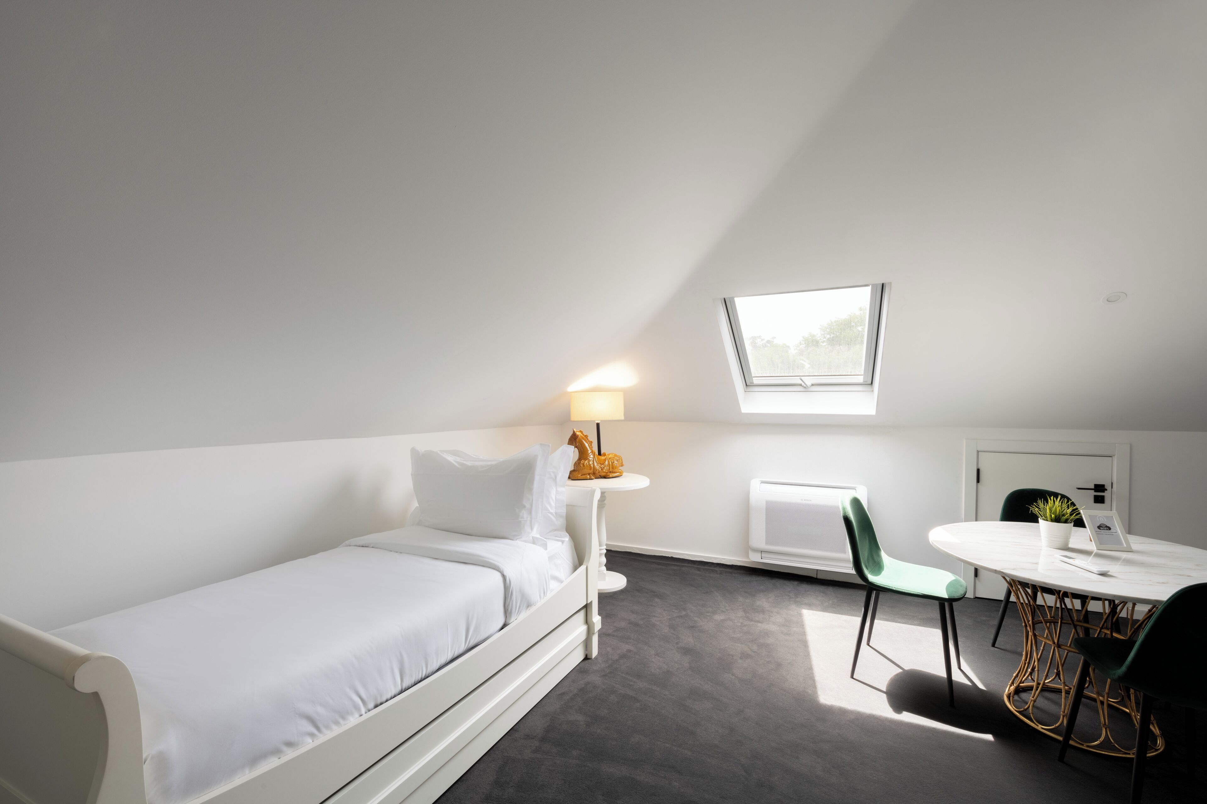 A modern, minimalistic room with a white bed, a small round table with a laptop, a green chair, and a window providing natural light.