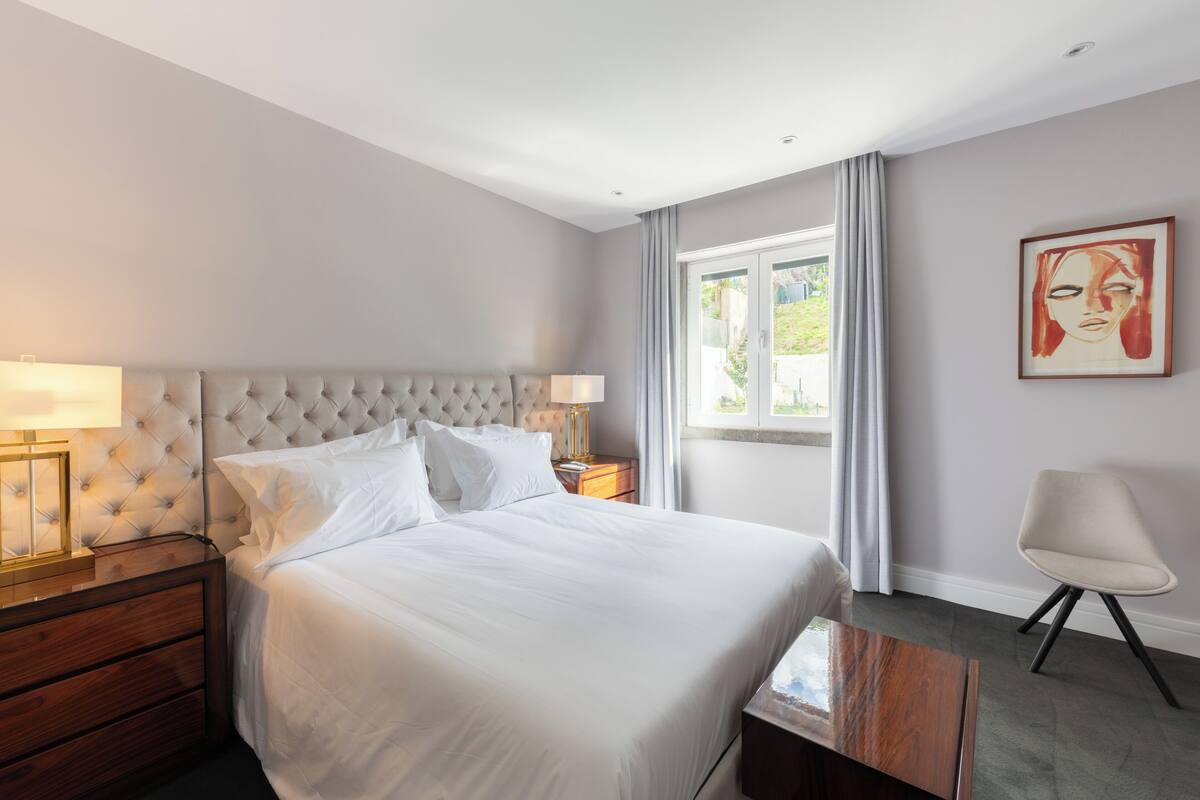 Ein modernes Hotelzimmer mit einem gepolsterten Bett, einem Nachttisch mit Lampe, einem Sessel und einem großen Fenster mit Vorhängen.
