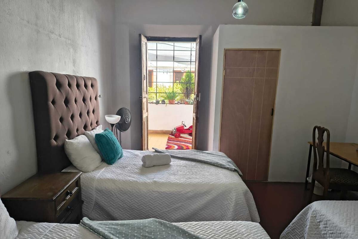 A bedroom with two beds, a desk, and a chair. There is a window with a view of a red car and greenery outside.