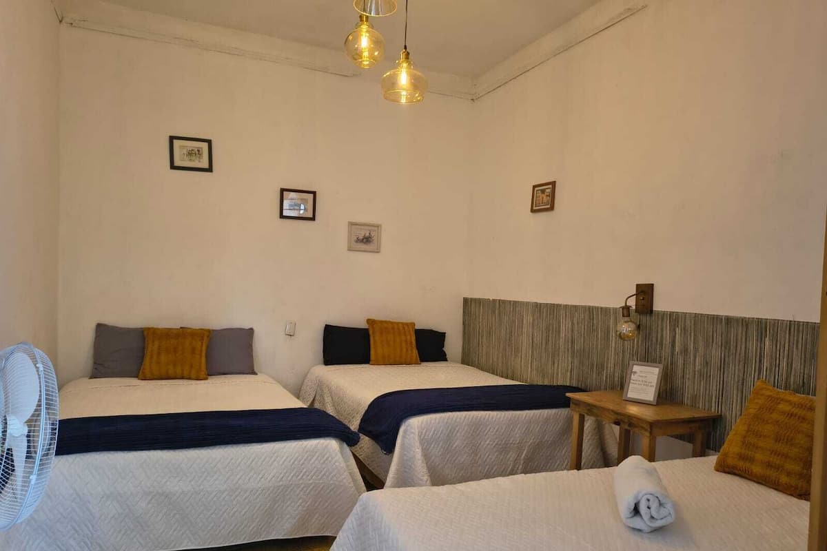 A hotel room with two beds, a small table, a fan, and wall-mounted lights.