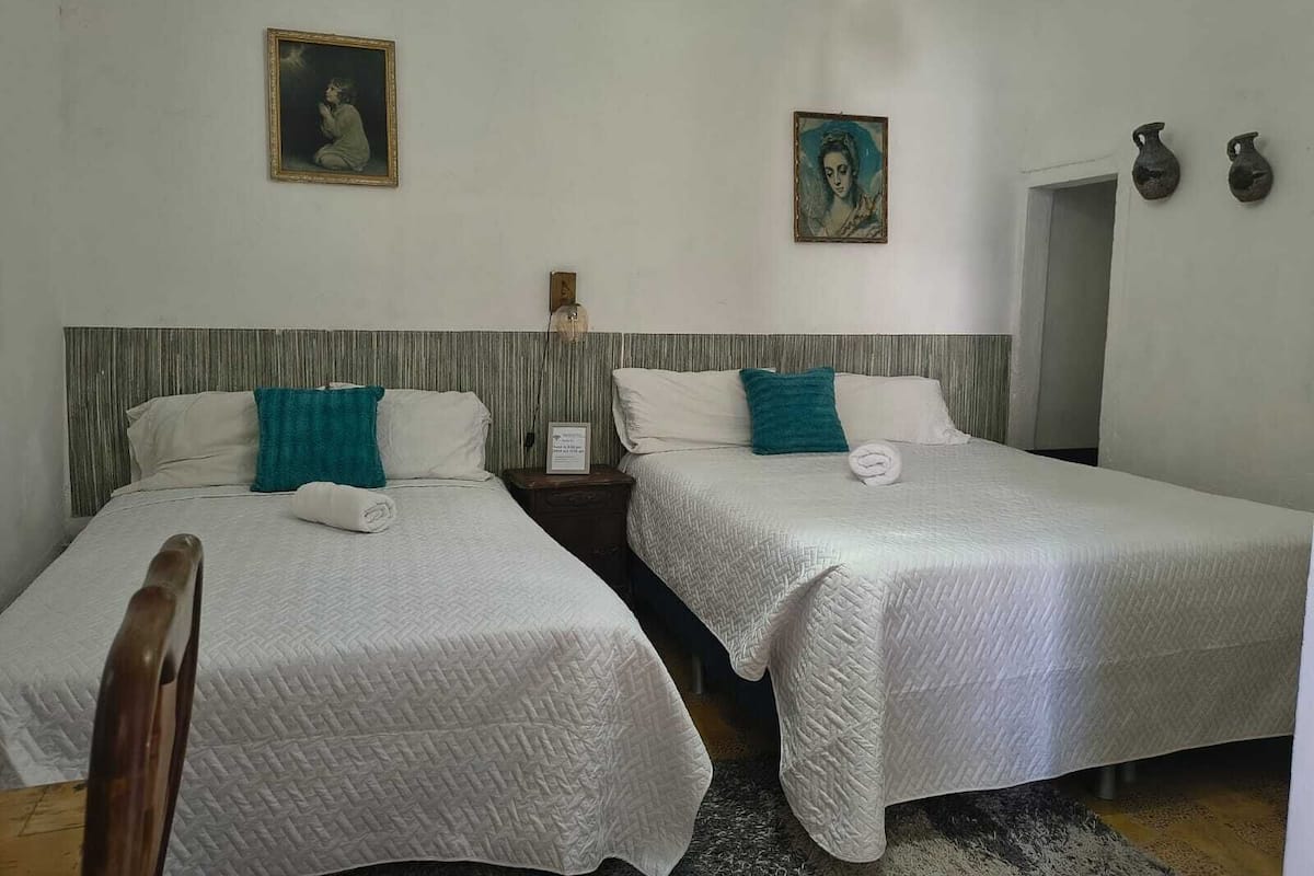 A hotel room with two beds, each with a white quilted bedspread and teal pillows. There are two paintings on the walls, a small clock, and a door with two vases on the wall.