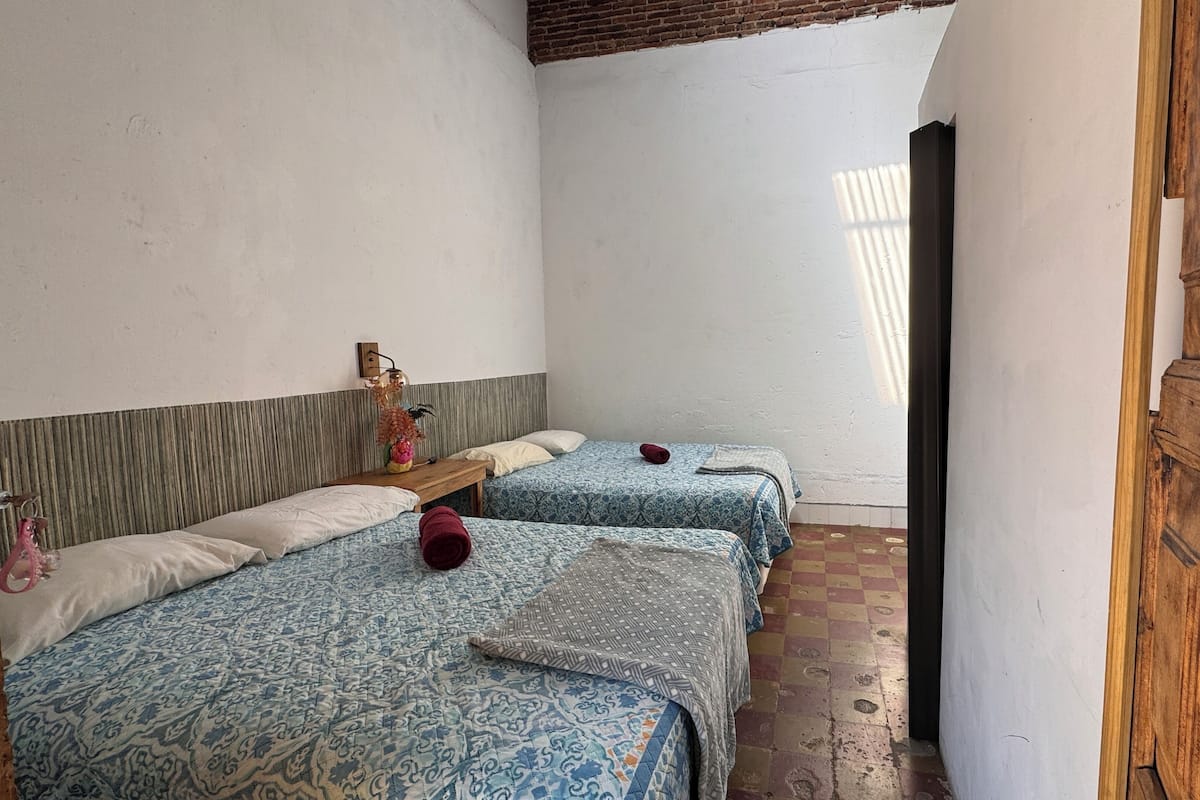 A narrow room with two beds, each with patterned bedding and a wooden headboard. A small table with a vase and a keychain is visible.