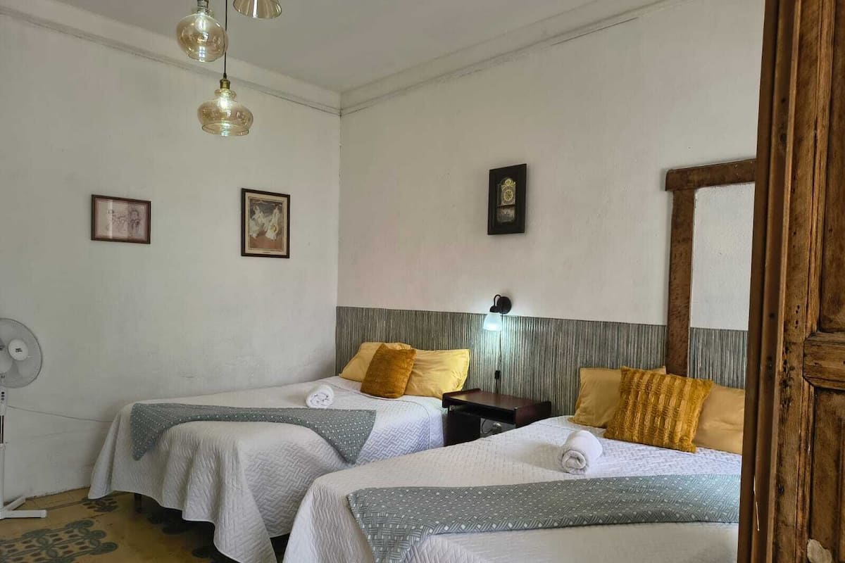 A hotel room with two beds, each with a white bedspread and a patterned blanket. There are wall-mounted lamps, a fan, and framed pictures on the walls.