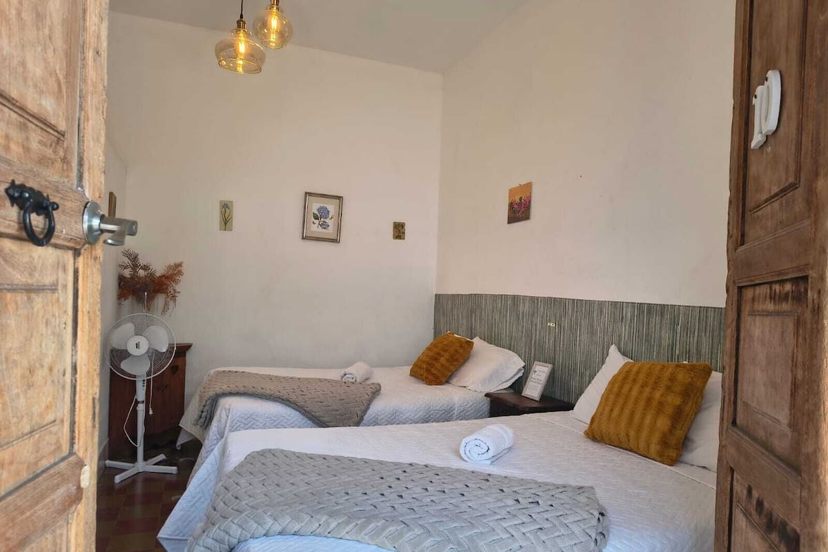 A room with two beds, a wooden door, a fan, and wall-mounted lights.