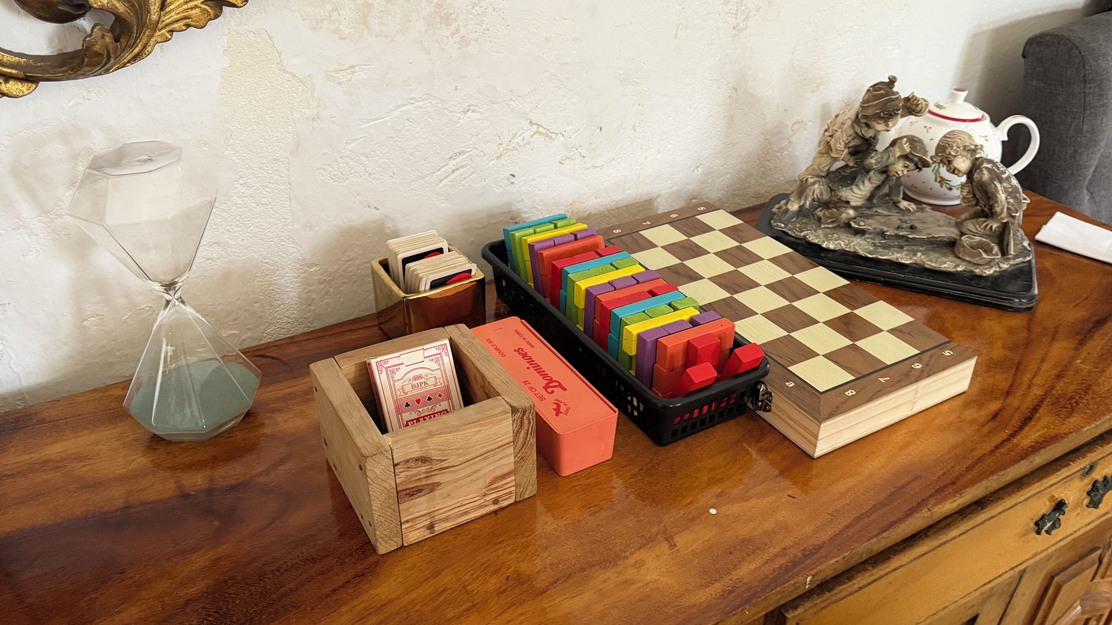A wooden table with a chessboard, colorful blocks, a glass hourglass, and a teapot.