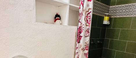 Un baño con inodoro, un cubo de basura, una repisa con un objeto decorativo, ducha con cortina estampada y un dispensador de papel higiénico.