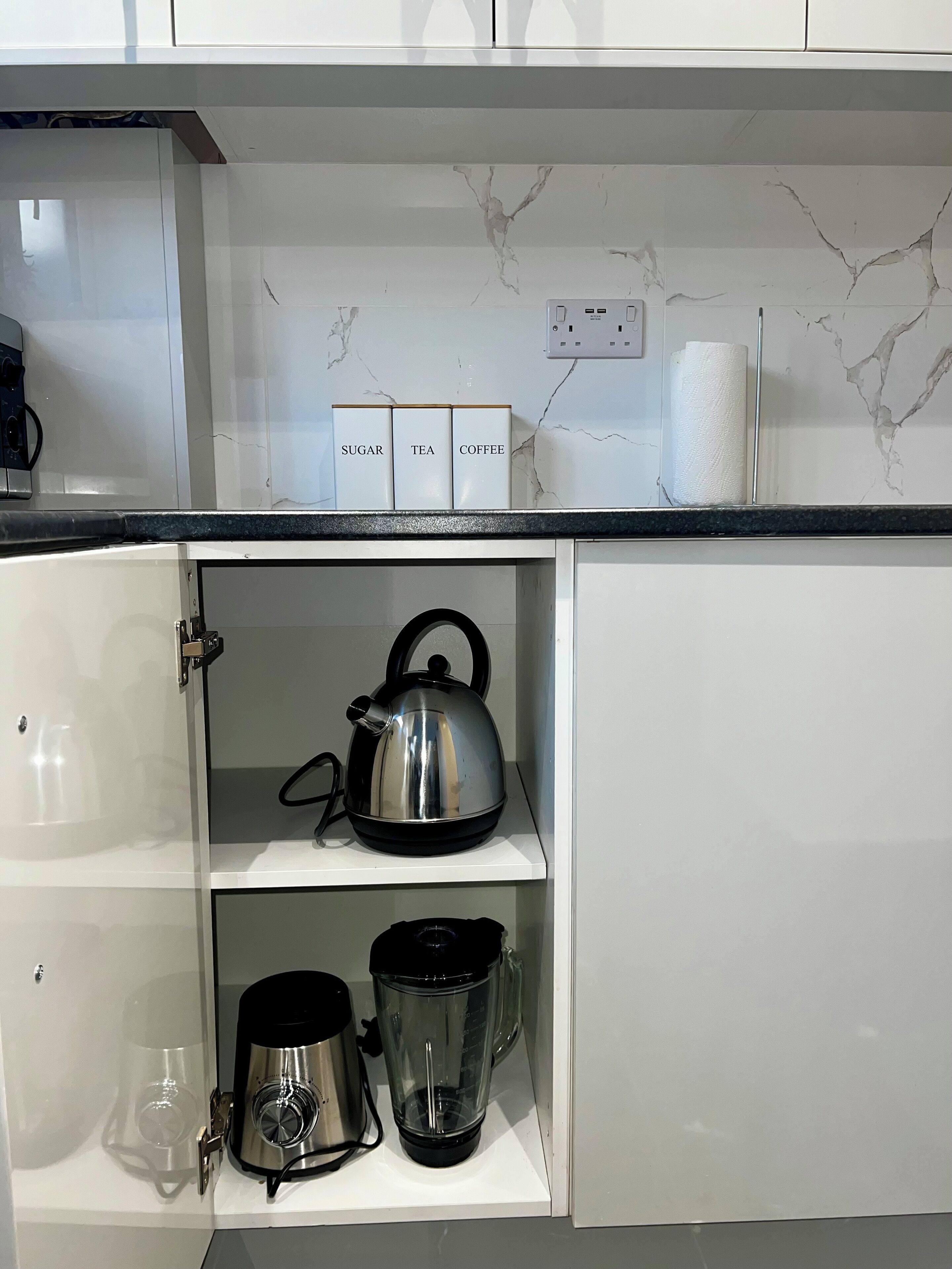 Una cocina con salpicadero de mármol, un hervidor, una cafetera y cajas etiquetadas para azúcar, té y café.