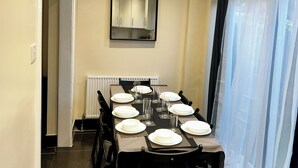 Un comedor con una larga mesa dispuesta para cuatro, sillas, un reloj en la pared, luces empotradas en el techo, una ventana con cortinas traslúcidas y un radiador debajo del espejo.