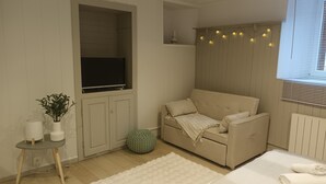 A cozy room with a beige sofa, a flat-screen TV, a small table with a plant, and a window with string lights.