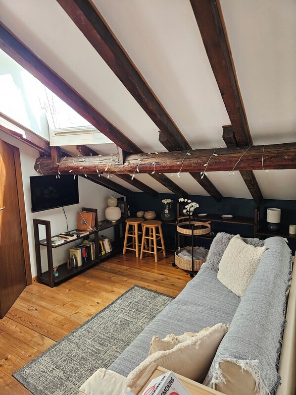 Een loft-achtige kamer met zichtbare houten balken, een bank, een kleintje tafeltje en een boekenkast.