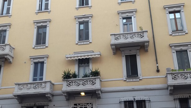 Un immeuble à plusieurs étages, doté d’une façade jaune, de nombreuses fenêtres avec volets et d’une entrée centrale ornée d’une grille métallique décorative.